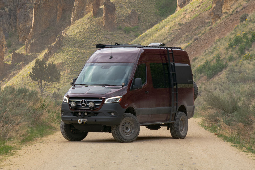 Red van on a dirt road, showcasing the OPTI-RATE STRIKER AWD 2 LIFT KIT for Sprinter AWD, enhancing vehicle's clearance and off-road capability with larger tires.