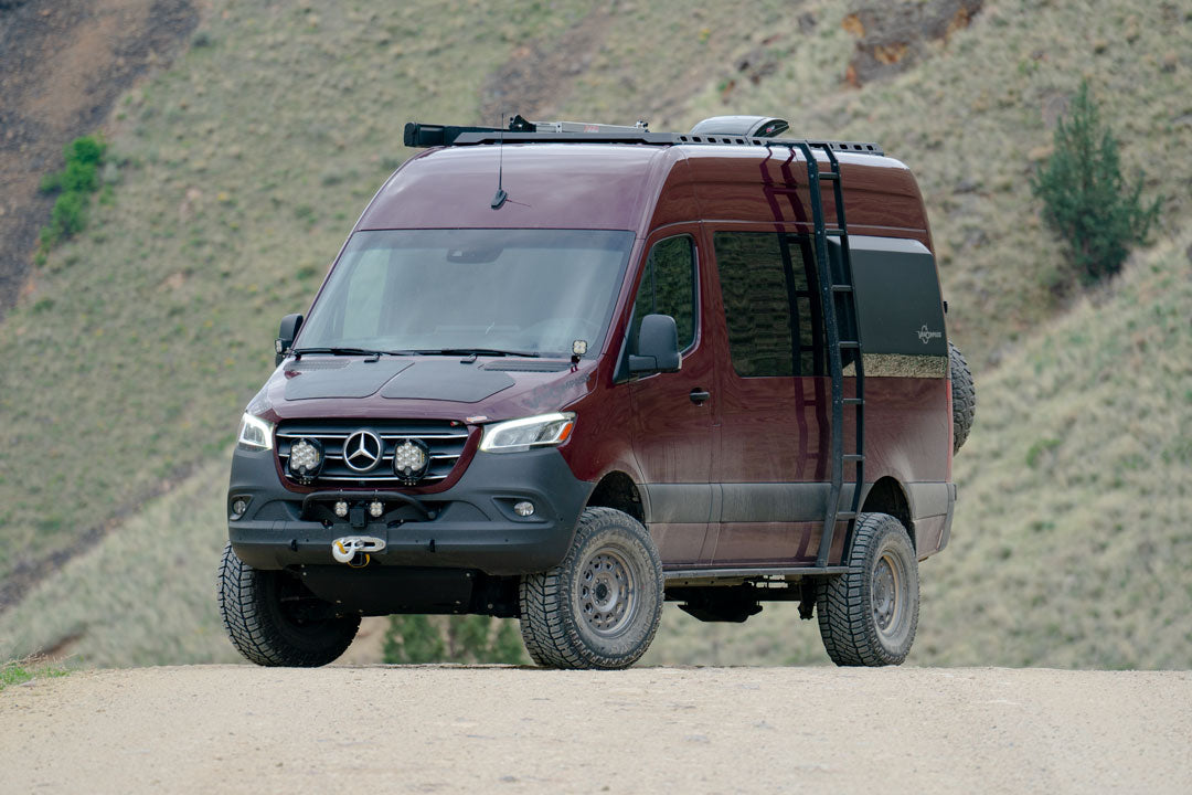 A red van with roof racks parked on a dirt road, with a visible close-up of its tires.