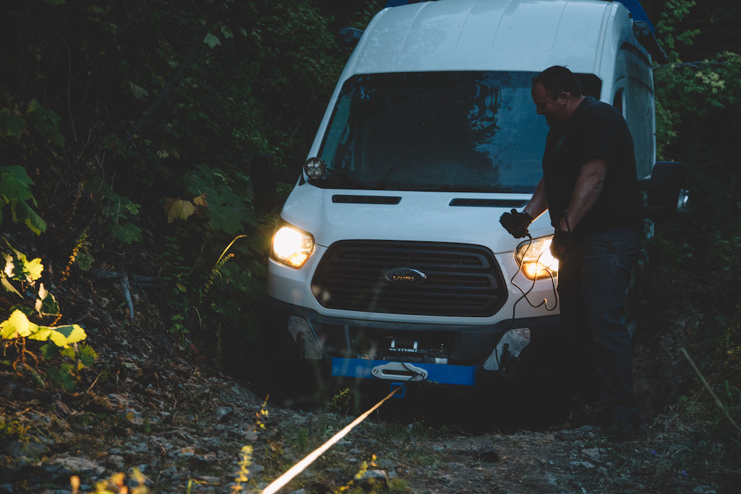A man standing next to a white van, holding a wire, with the vehicle's headlight and vent visible.
