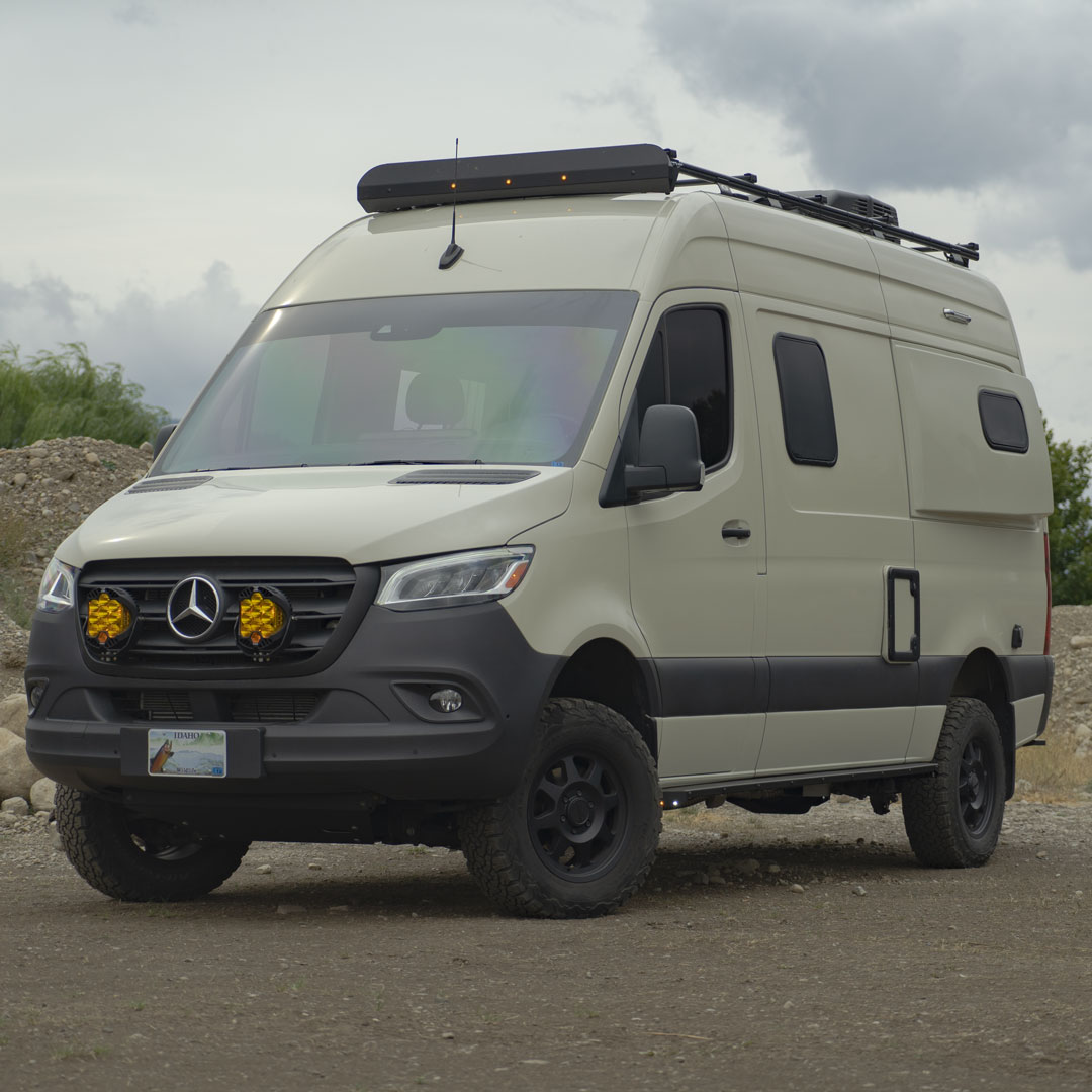 A white van parked on a rocky surface with visible close-ups of its black tire, yellow light, logo, and headlight.