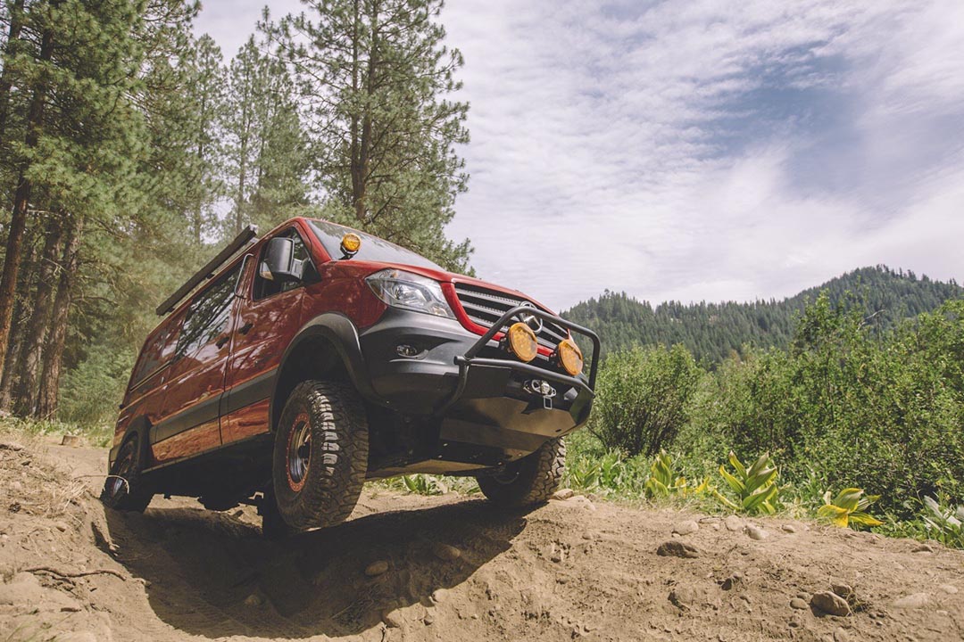 A red truck off-roading on a dirt hill with a forest backdrop, featuring visible tires and a side view mirror.