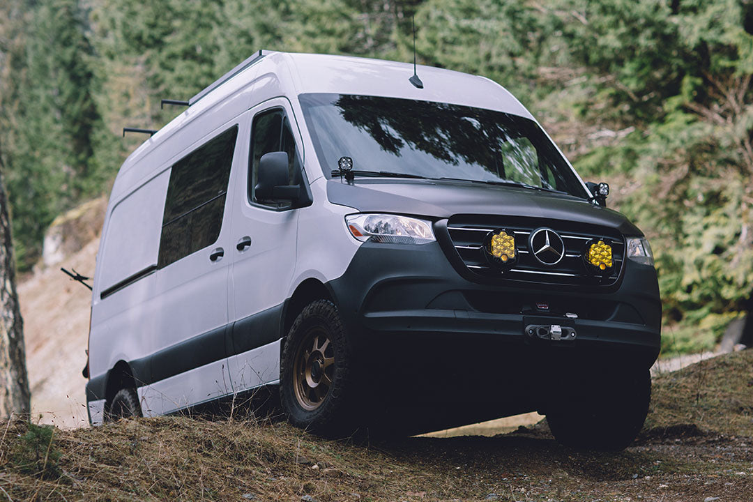 A white van parked on a dirt road with visible close-ups of its tire, yellow light, and rearview mirror.