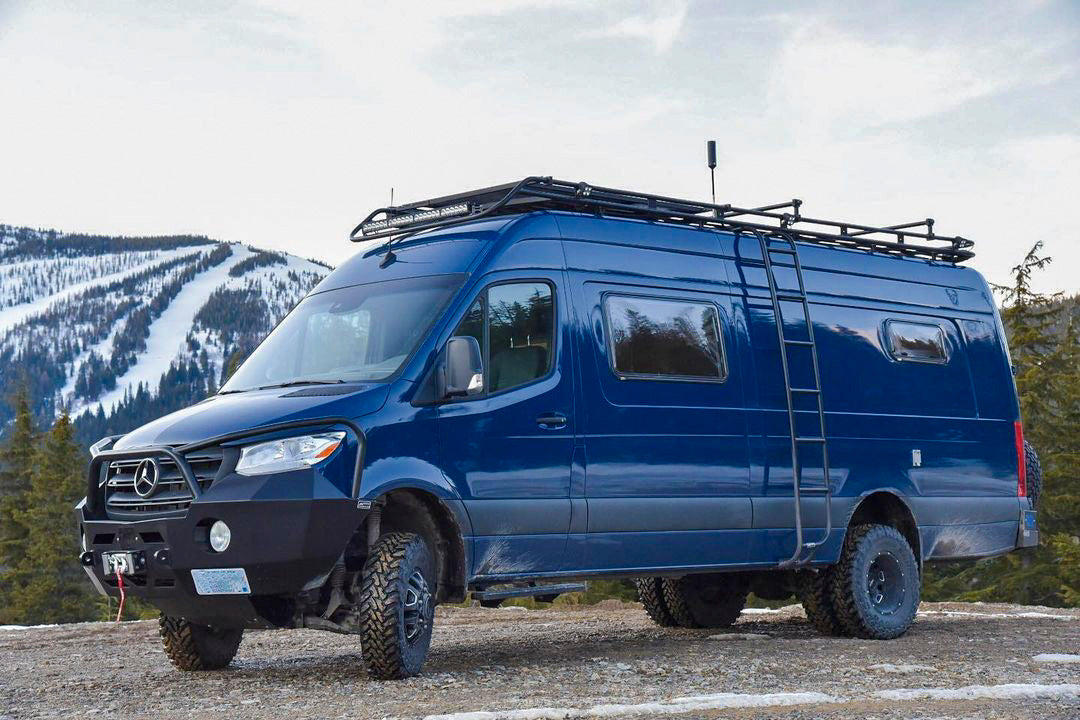 A blue van parked on a gravel road with mountains in the background. The image shows a side view with visible tires and a headlight.