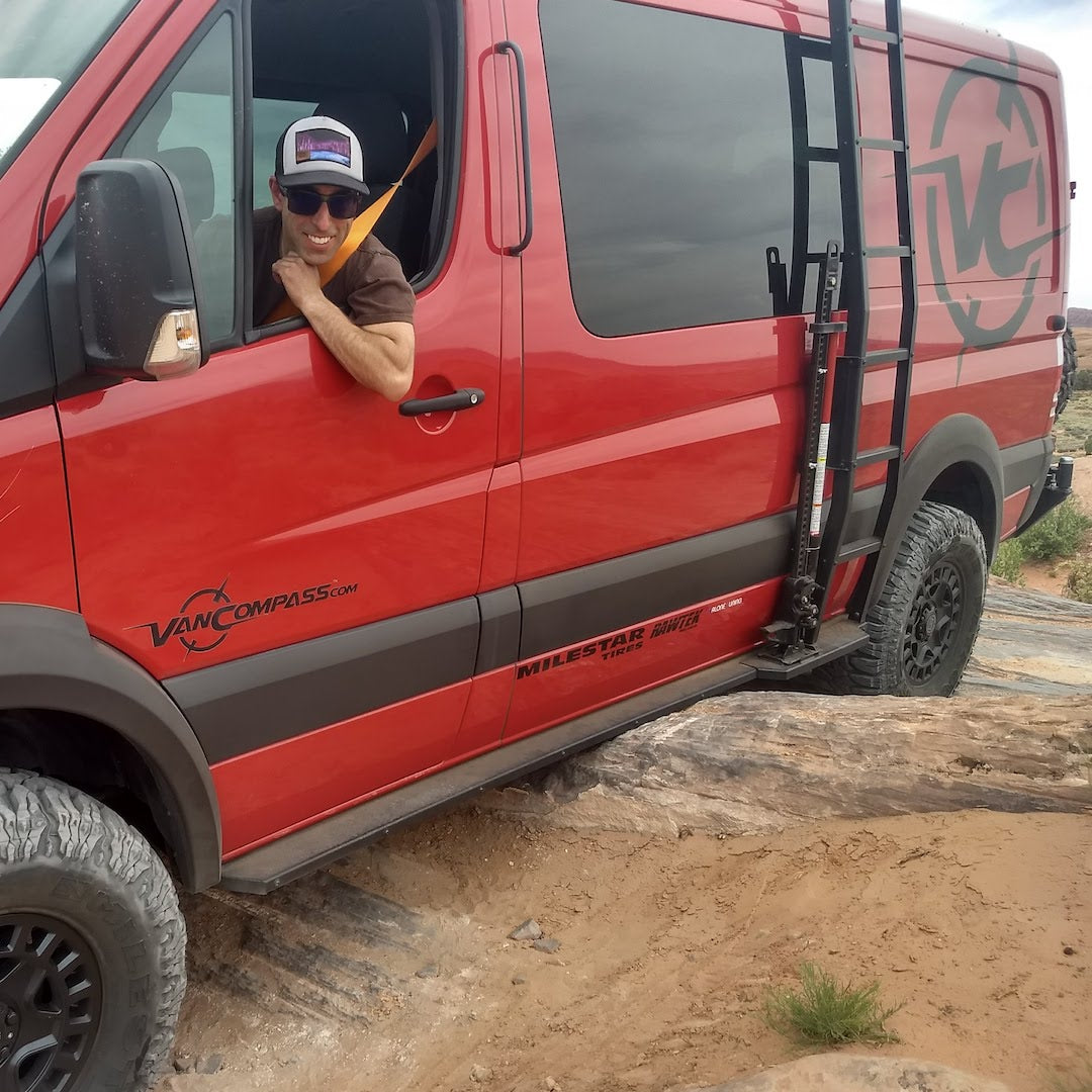 Man leaning out of red van window showcasing ROCKER GUARDS - SPRINTER 4X4 (2015-2022 170 WHEELBASE ONLY) by VAN COMPASS, visible tire and vehicle parts.