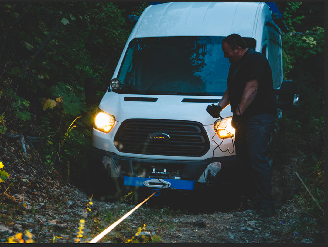 Man standing next to a white van, holding a WARN VR EVO 12S - 12,000LB WINCH featuring a synthetic line and relocatable control pack for heavy-duty pulls.